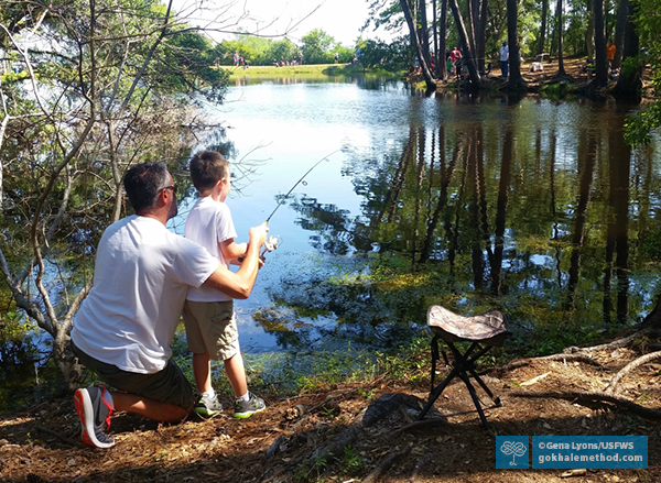 Photo of a man kneeling with his son, teaching him to fish at a lake.