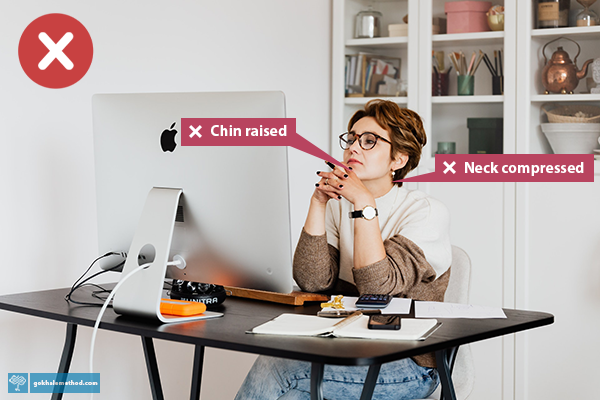 Woman at desk using computer, body rounded and chin lifted to focus on screen through glasses.